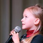 Cute young girl passionately singing into a microphone on stage, captured in a close-up profile view.